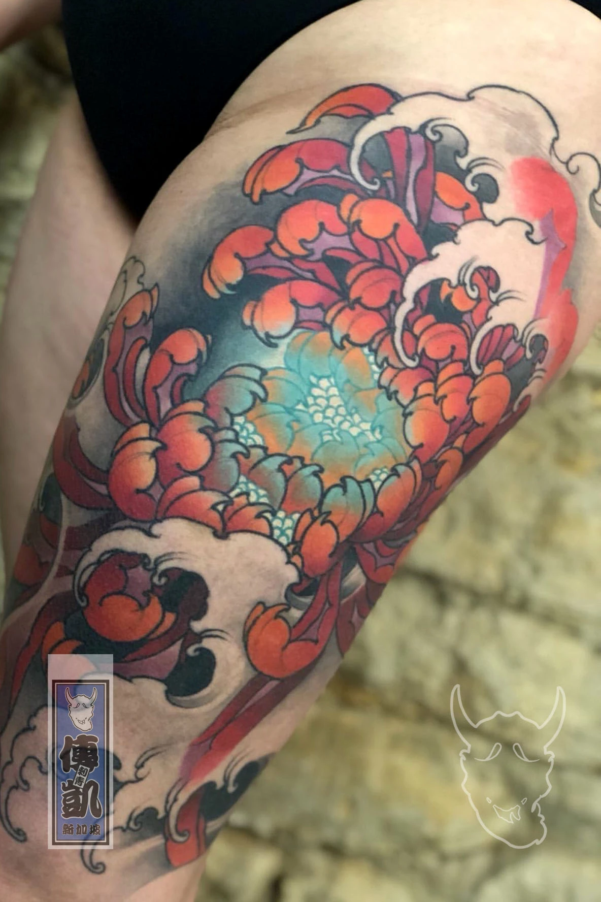 Traditional Japanese chrysanthemum (Kiku) tattoo on the thigh with vivid orange, red, and teal petals surrounded by black and grey wind patterns. Artwork by CK Tattoo Singapore.