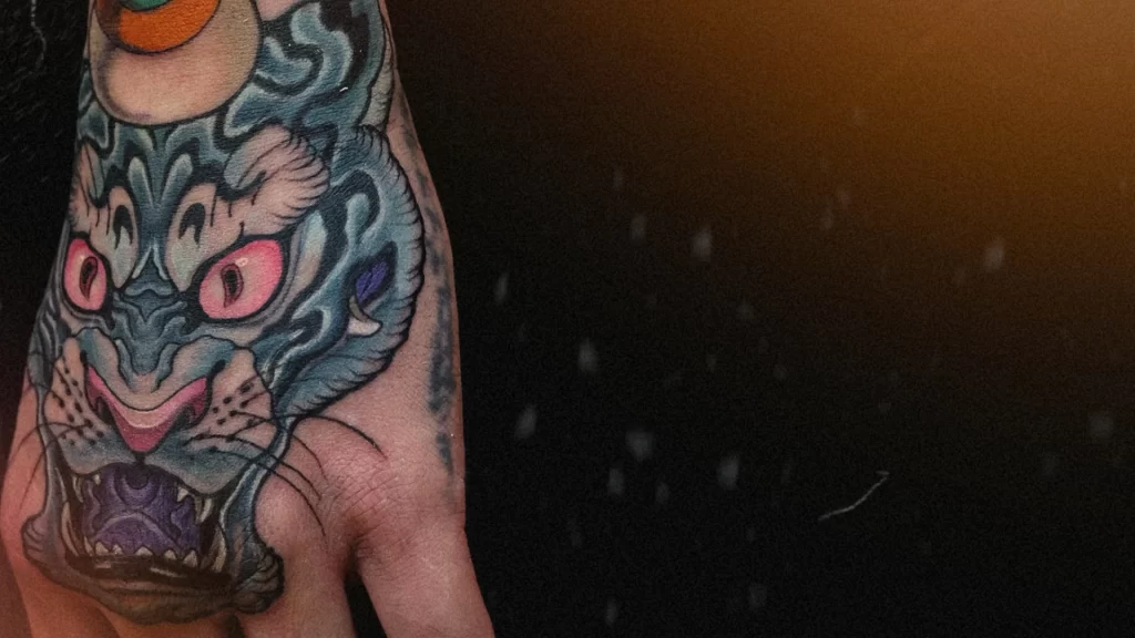 Traditional Japanese tiger tattoo on the hand, featuring blue fur, pink eyes, and an orange jewel above the head, symbolizing protection and strength. Artwork by CK Tattoo Singapore.