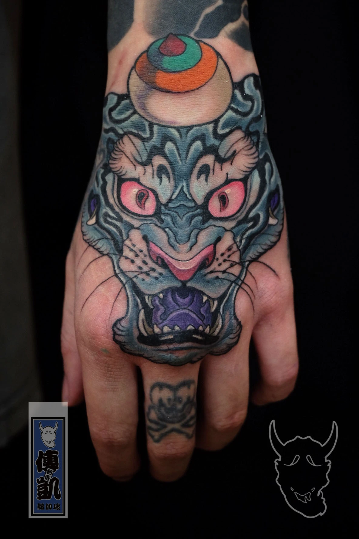 Traditional Japanese tiger tattoo on the hand, featuring blue fur, pink eyes, and an orange jewel above the head, symbolizing protection and strength. Artwork by CK Tattoo Singapore.